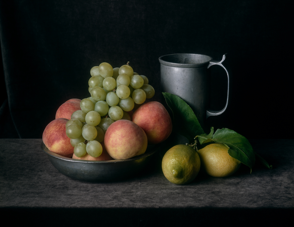 Colour photograph. A bowl filled with apples and grapes sits on a table beside a pitcher and two lemons.