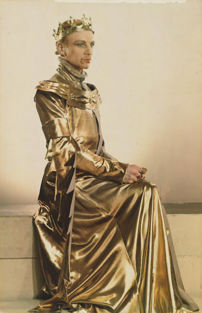 Colour photograph. John sits in profile, wearing a long golden gown and a crown.