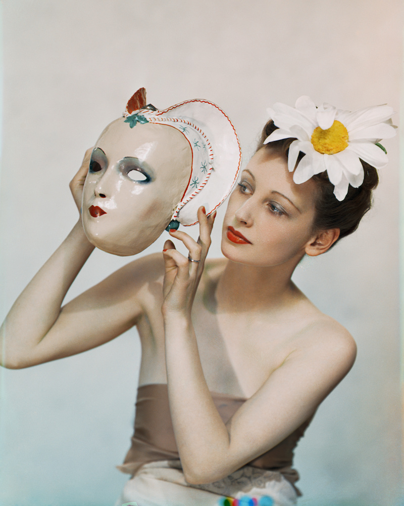 Colour photograph. Rosemary holds a mask aloft, which - decorated with blue eyeshadow and red lipstick - resembles her own make up. She wears a large fake daisy in her hair.