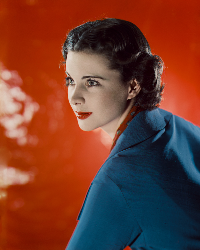 Colour photograph. Vivien Leigh wears a blue jacket and poses in front of a red background.
