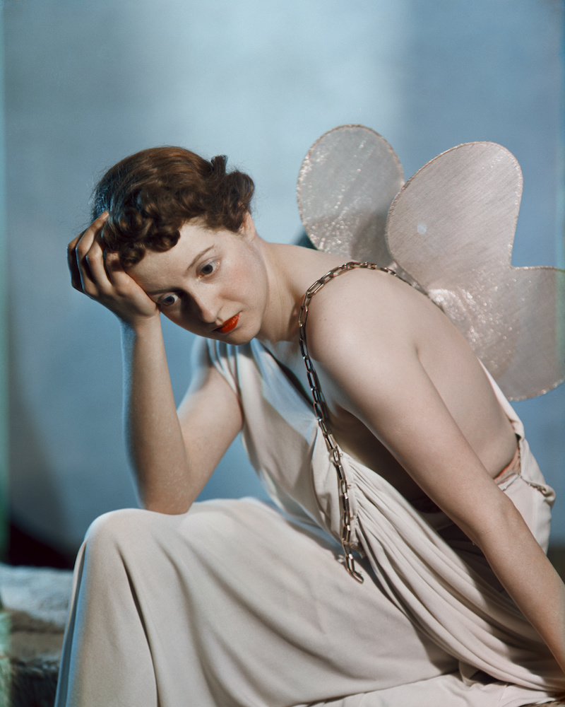 Colour photograph. A woman dressed in a white robe and white butterfly wings poses with her head resting on her hand. A golden chain hangs over her shoulder.