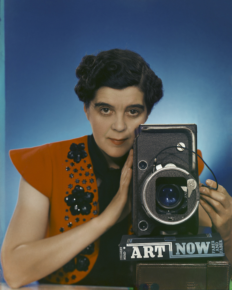 Colour photograph. Yevonde holds a large camera, which rests upon a book - 'Art Now', published by Faber and Faber. She wears a red jacket patterned with black flowers, and stands in front of a blue background.