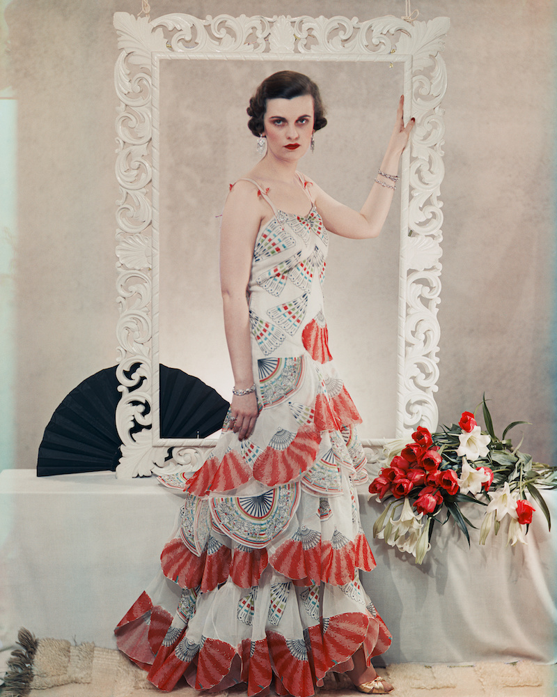 Colour photograph. A woman wearing a white dress decorated with a red fan pattern stands in front of a large white frame, which her hand rests upon. There is a black fan on the white table behind her, as well as a bouquet of red roses and white lilies.