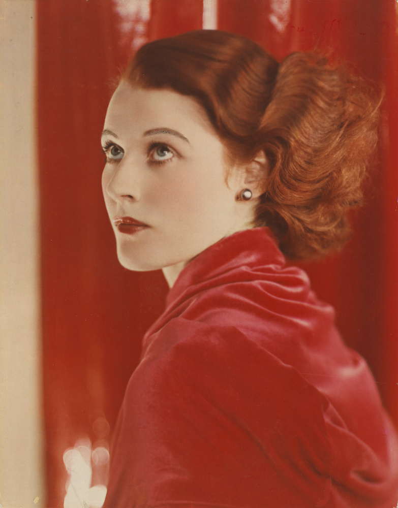 Colour photograph. Joan wears red and poses in front of a red background. We see her head and shoulders; she looks slightly upwards.
