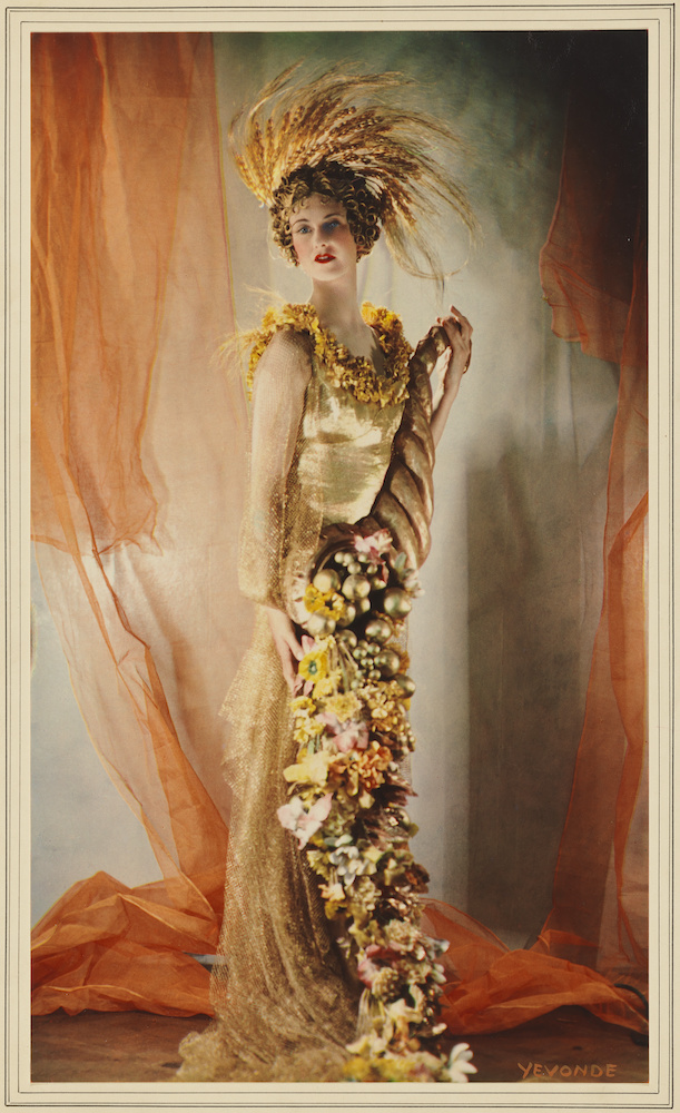 Colour photograph. Lady Dorothy poses in front of orange drapes, wearing a golden dress adorned with autumnal coloured flowers and golden berries.
