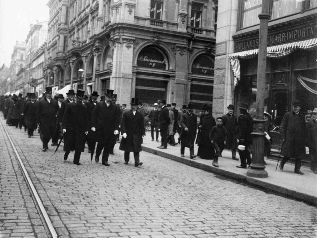 Black and white photo. Men in top hats and suits walk down a cobbled street.