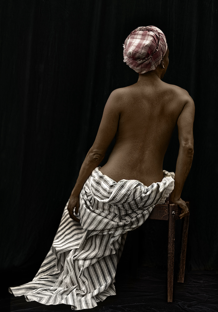 Colour photo. A Black woman wearing a red and white fabric hat and a black and white striped skirt reclines on a stool. Her back faces towards the camera.