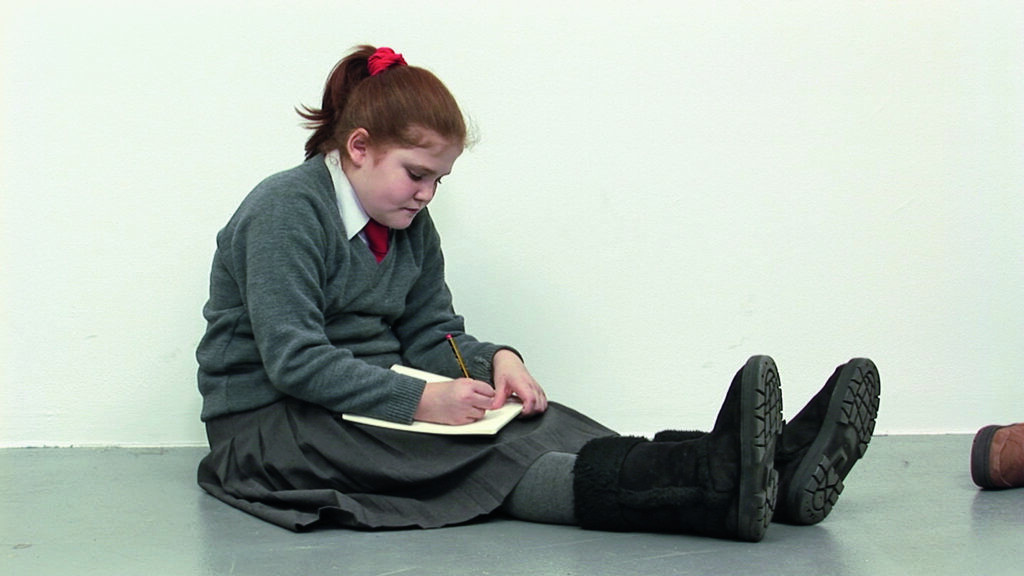 Film still. A young girl is sitting on the ground, drawing on the piece of paper on her lap.