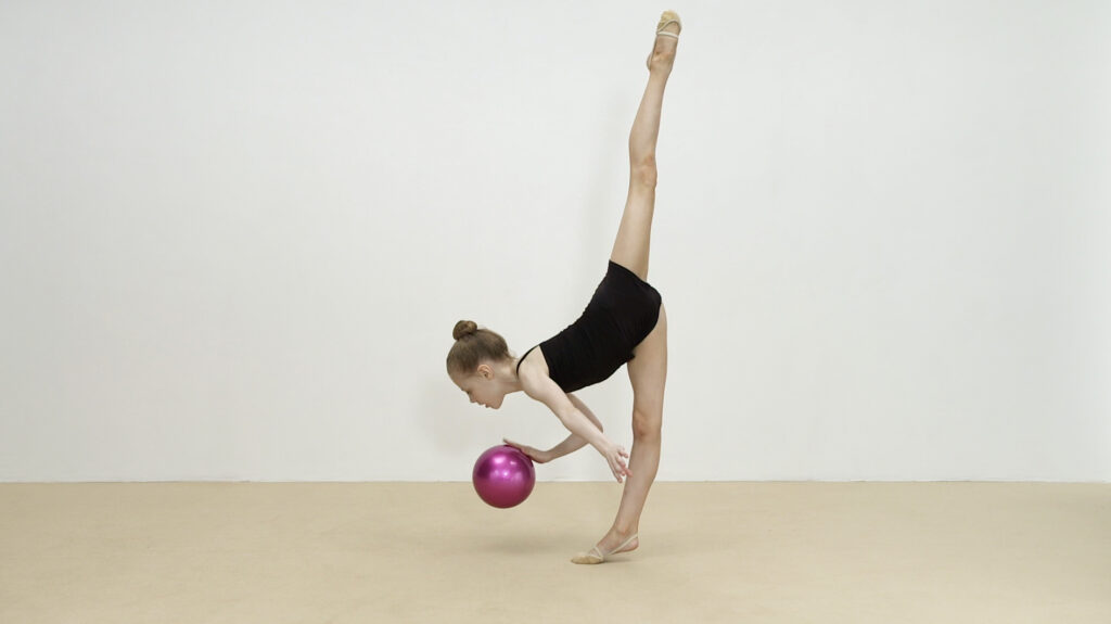 Film still. A young girl holds a gymnastics pose. She balances on one leg, her other leg straight up in the air. She holds a pink ball against one hand.