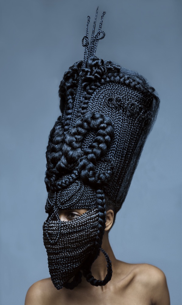 Colour photograph. A woman wears a towering, elaborate headdress comprised of braids of hair.