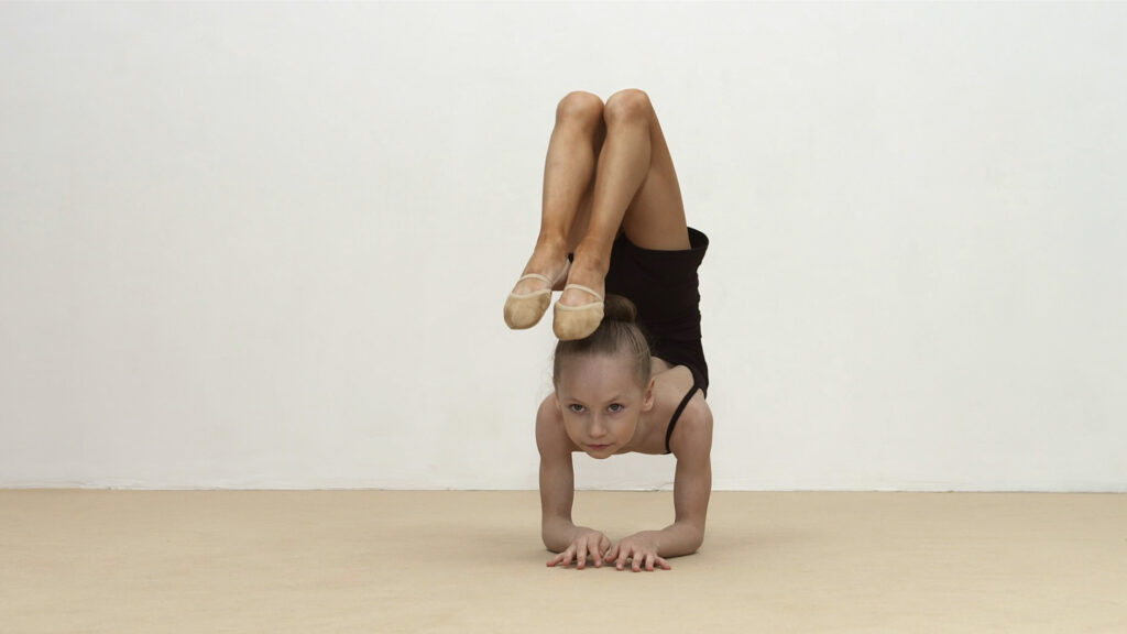 Film still. a young girl holds a gymnastics pose. She balanced on her forearms, her legs upwards, bending over her body so that her feet are above her head.