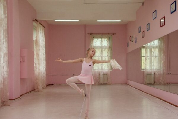 Film still. A young girl holds a ballet pose, balancing on the point of one foot, in a ballet studio. The studio is painted pink, and she wears a pink dress, tights and shoes.