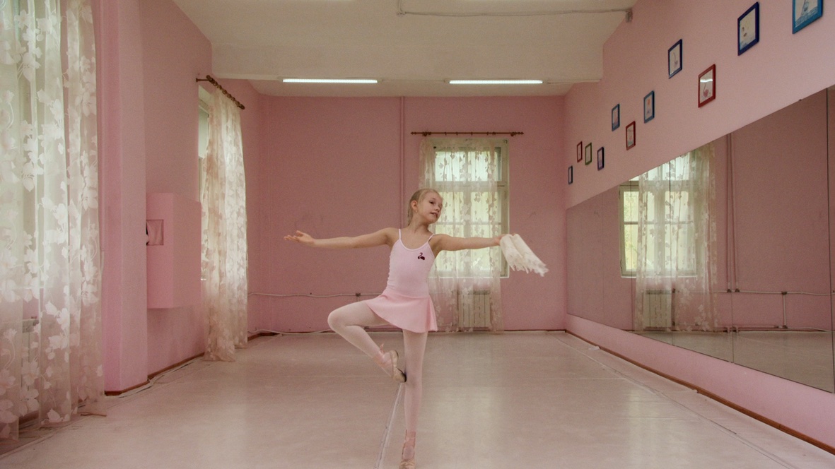 Film still. A young girl holds a ballet pose, balancing on the point of one foot, in a ballet studio. The studio is painted pink, and she wears a pink dress, tights and shoes.