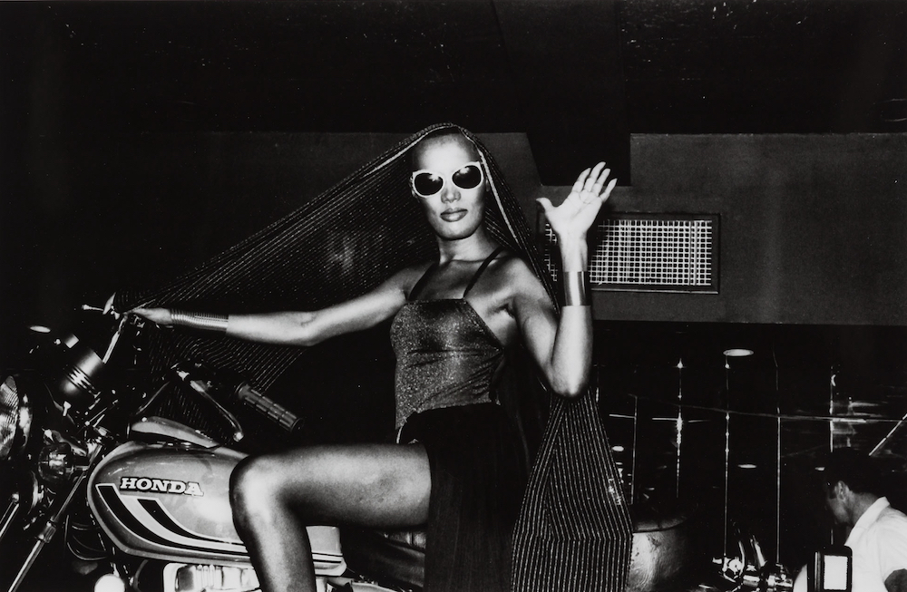 Black and white photo of Grace on a Honda motorcycle. They wear sunglasses and wave to the camera.