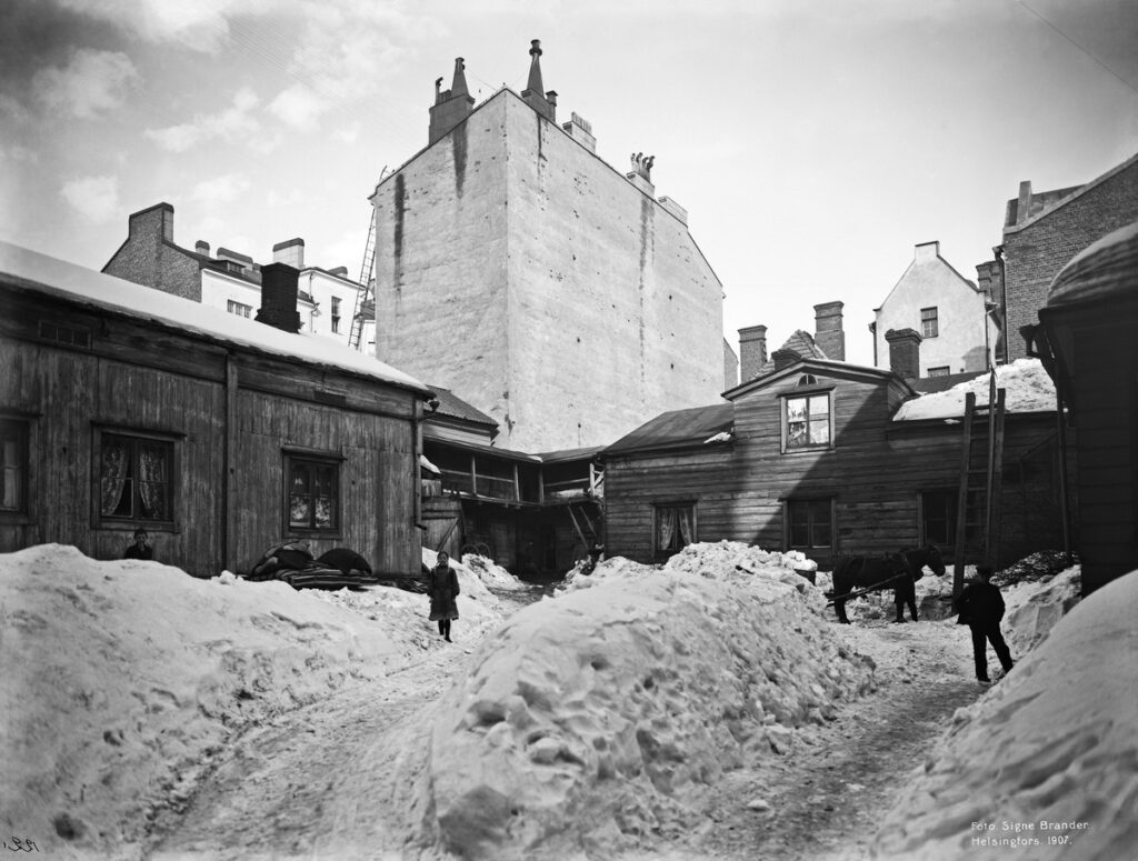 Black and white photo. Small wooden buildings are dwarfed by an imposing brick building behind them. In the foreground, people and a horse walk in snow.
