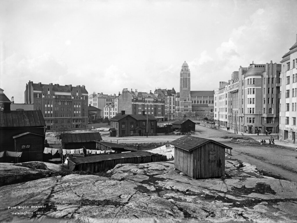 Black and white photo of Helsinki in 1912. Wooden buildings stand in front of a larger expanse of buildings, constructed with bricks.