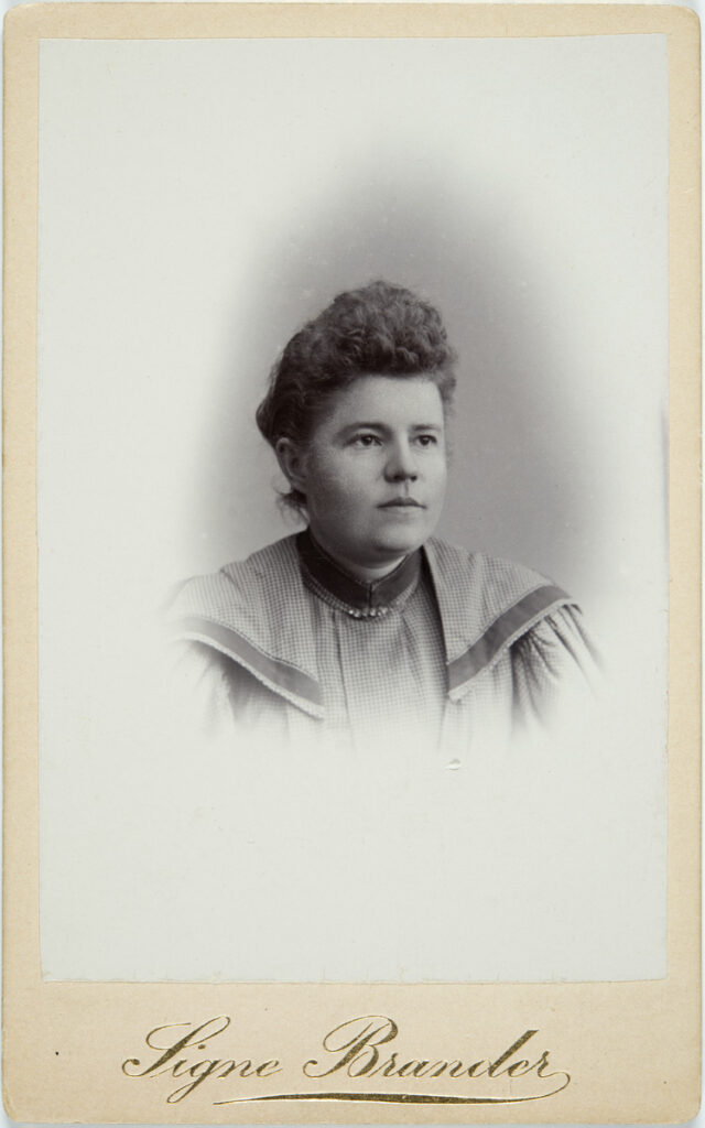 Black and white self portrait of Signe Brander. She wears a gingham dress with a collar.