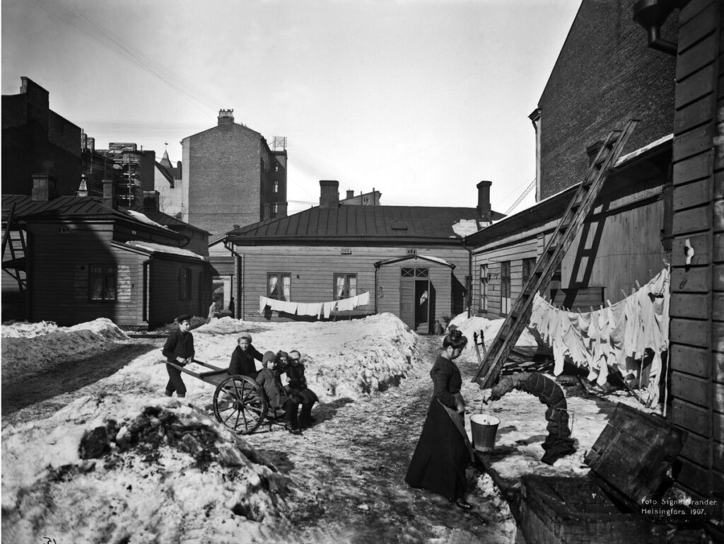 Black and white photo. A woman walks while a group of young children sit on a cart, pushed by a young boy. There are wood panelled and brick buildings, washing hung on lines, and the ground is covered with mounds of snow.