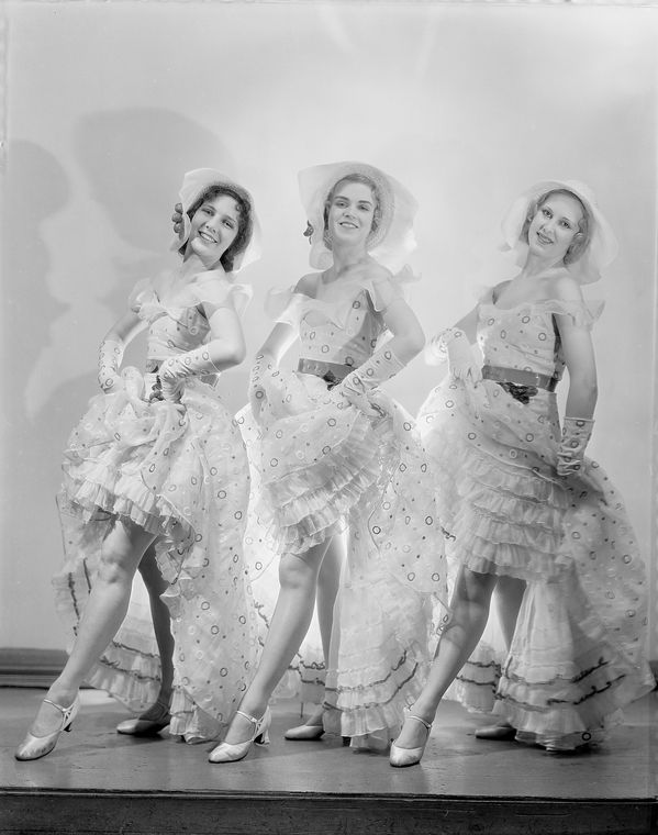 Black and white photograph. Three women wearing ruffled dresses and hats, posed with one leg in front of the other.