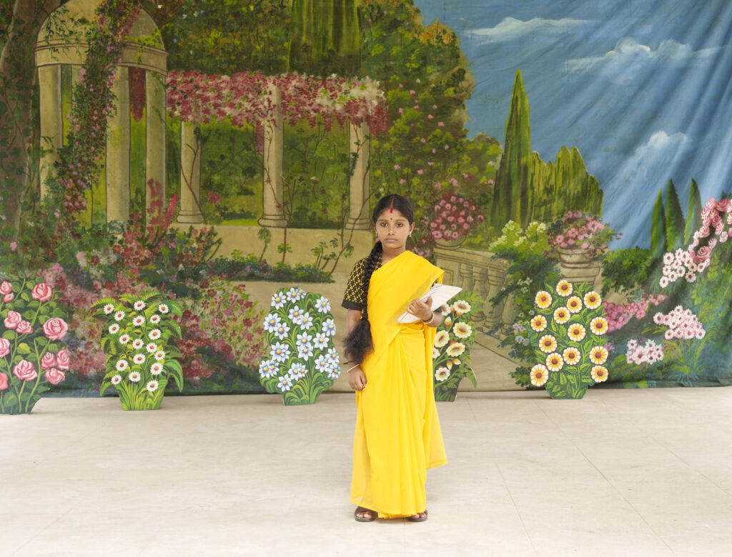 Colour photograph. A young girl in a yellow sari is holding a notebook. She is standing in front of a painting of a flower garden.