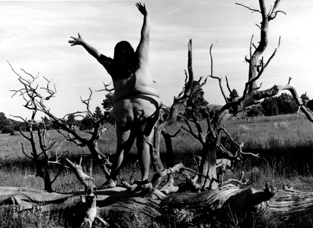 A woman stands naked, facing away from the camera and throwing her arms into the air. She is standing on a log with branches forking in different directions.