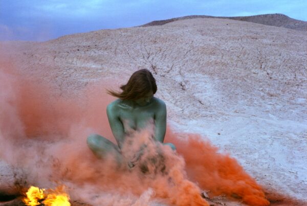 A woman, painted in blue, sits amid plumes of orange smoke and small fires, on a dry landscape.