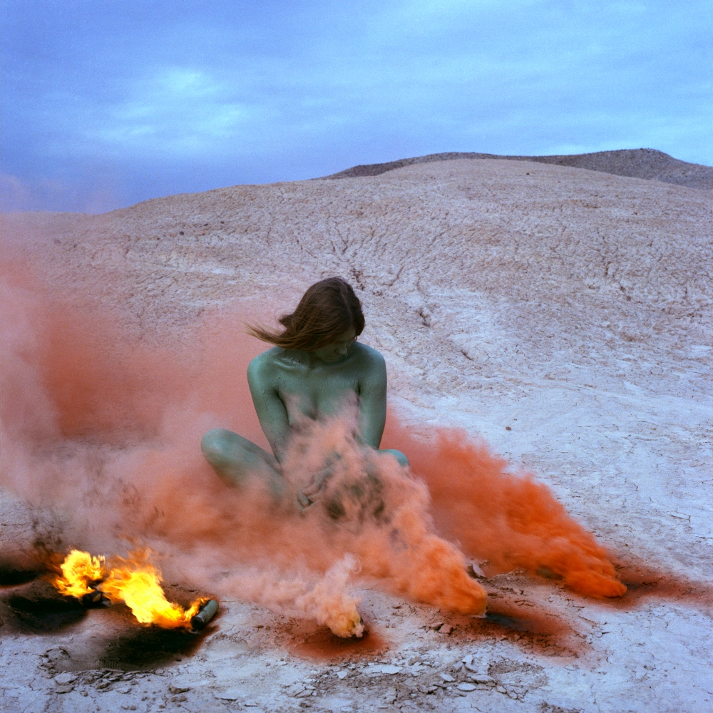 A woman, painted in blue, sits amid plumes of orange smoke and small fires, on a dry landscape.