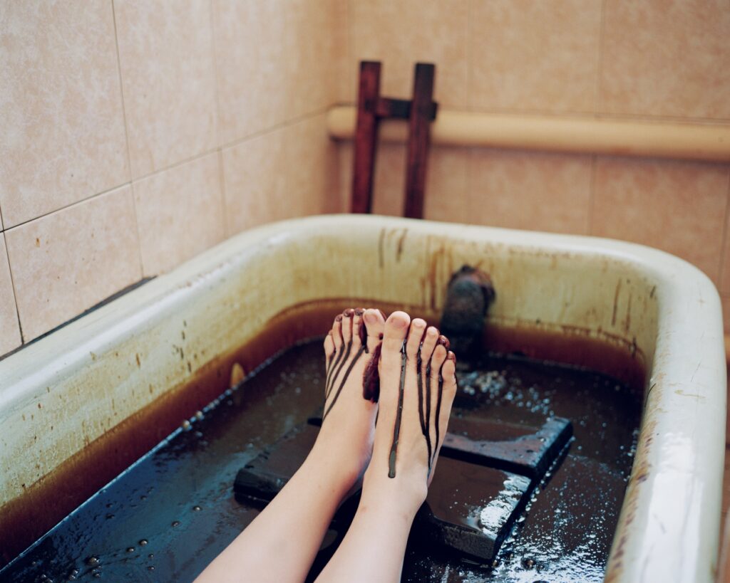 Colour photograph of a person bathing in oil. Only their feet and calves are visible.