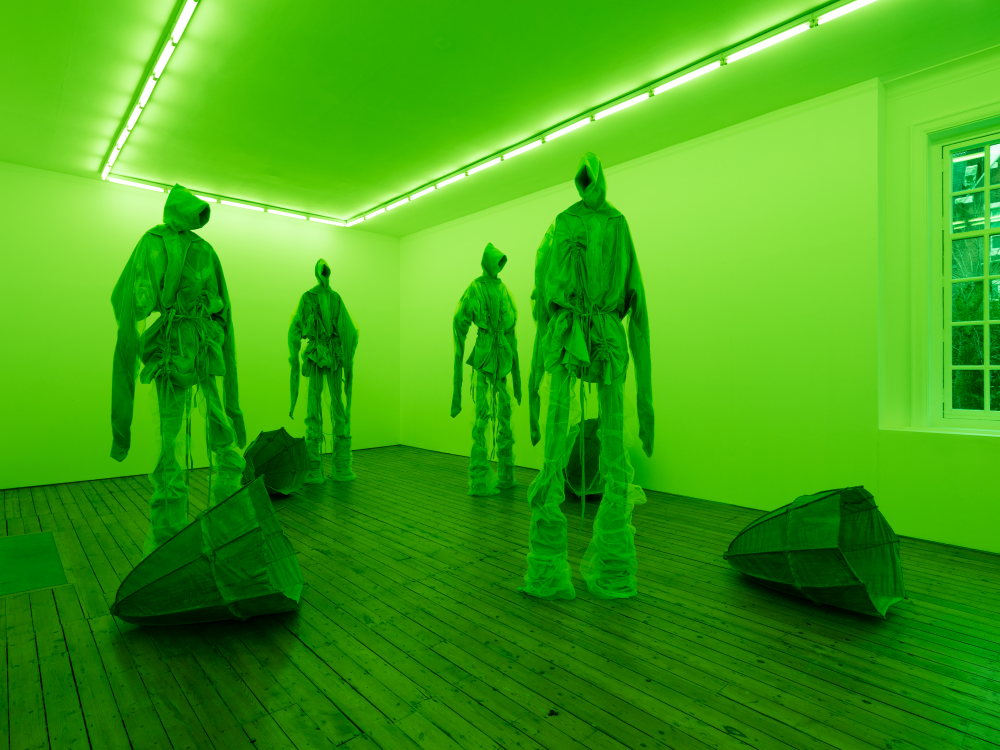 Four sculptures the the shape of humanoid figures stand in a room lit by bright green lighting. Four sculptural 'baskets' lie on the wooden panelled floor.