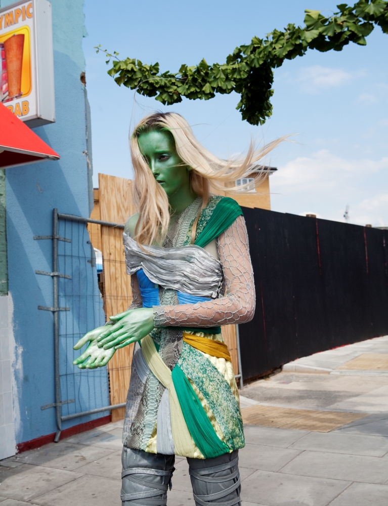 A blonde haired woman walks down a street. Her skin is painted green. She wears grey trousers and a long top made of green, yellow, and grey fabric.