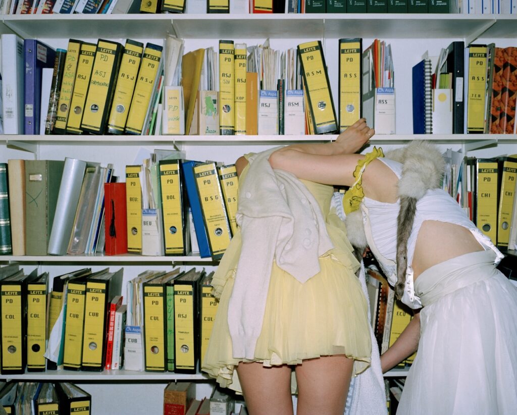 Two women in yellow and white dresses lean forwards towards a bookshelf, filled with folders with labelled yellow spines. We cannot see their heads as they reach into the shelves.