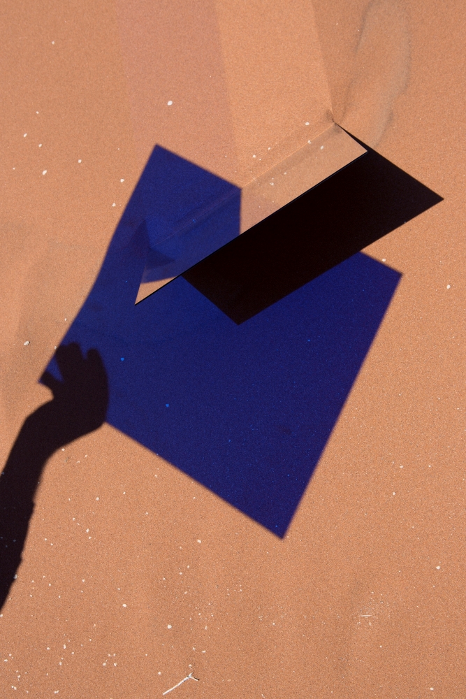 A blue. square shaped shadow is reflected onto a sandy floor. A silhouettes hand reaches towards it.