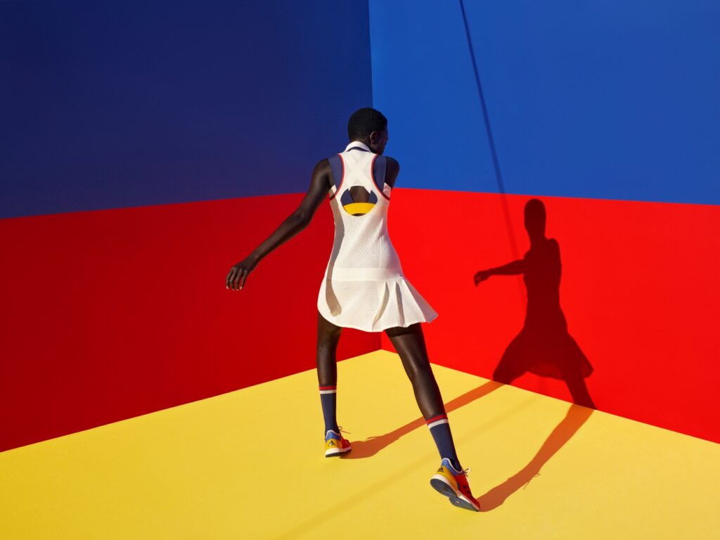 A woman in a white sports dress and Adidas shoes faces away from the camera, towards a blue and red striped wall and a yellow floor.
