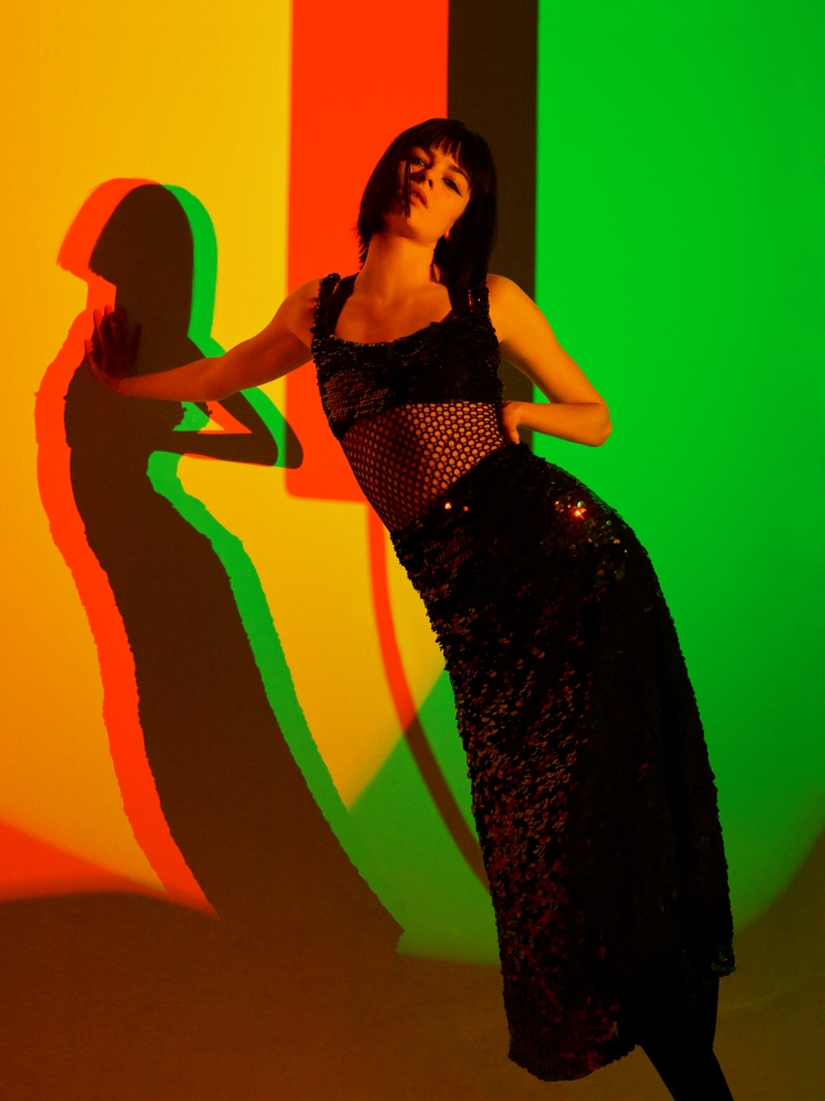 A woman wearing a black mesh top and a long, sequined black skirt leans against a yellow, orange, and green background.