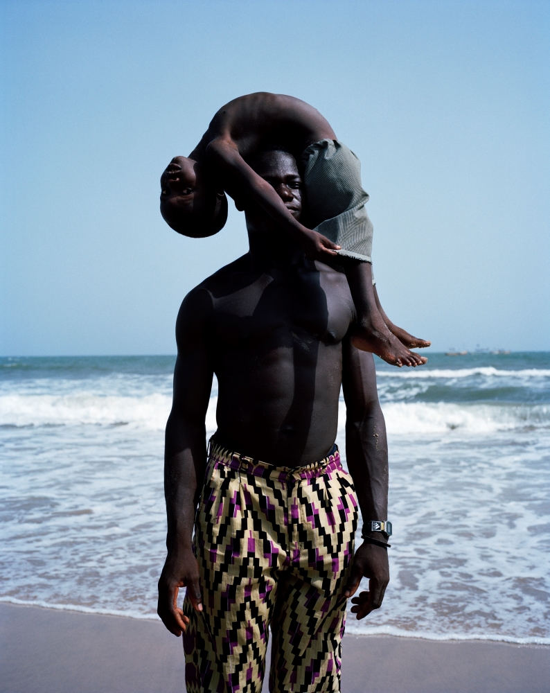 A man stands in front of the sea, on a beach. A young boy is draped over his head in a balanced pose.