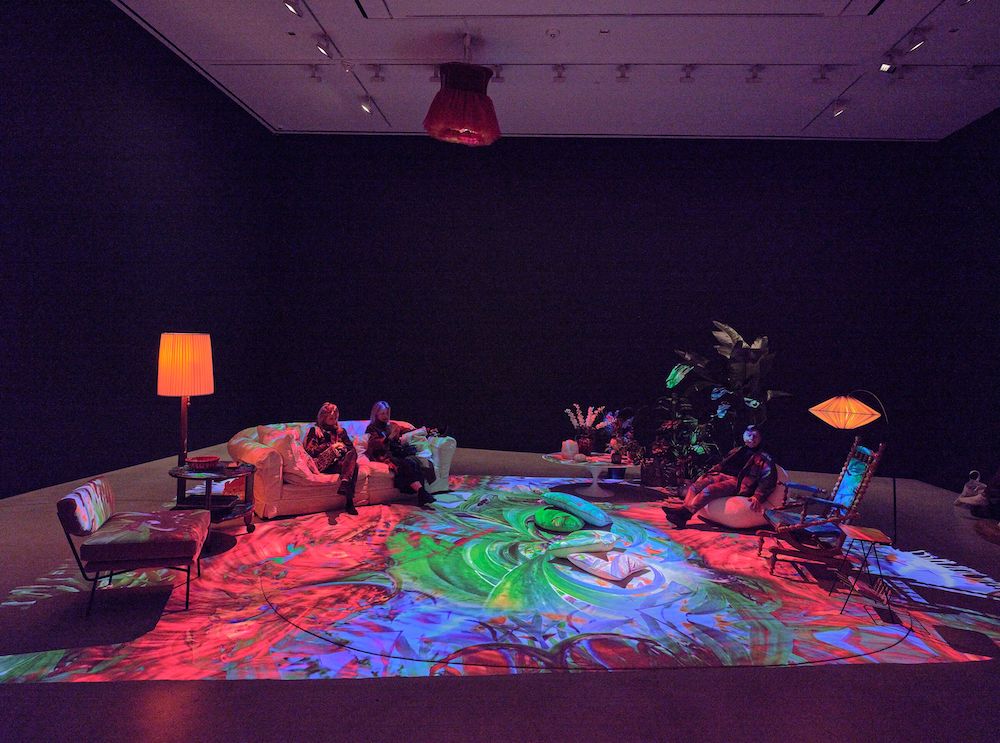 Installation view showing a 'living room' set up - sofas, chairs, coffee tables, and lamps - beneath swirling red, green, and blue lights.
