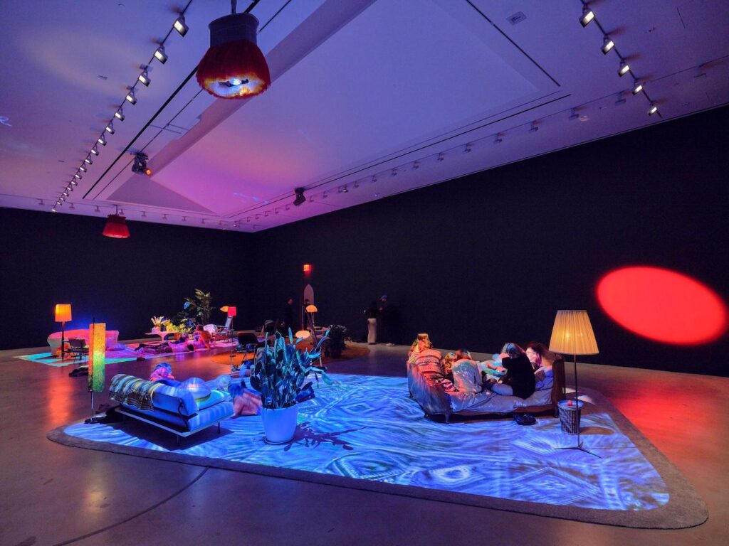 Installation view showing a 'living room' set up - sofas, chairs, houseplants, and lamps - beneath swirling pink and blue lights.
