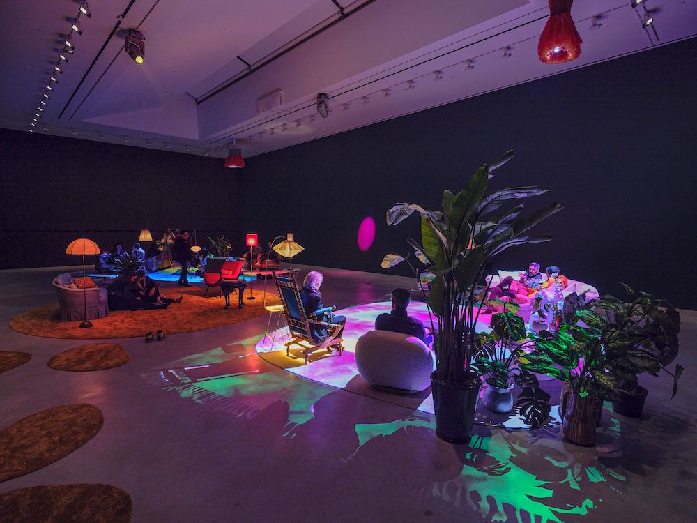 Installation view showing a 'living room' set up - sofas, chairs, houseplants, and lamps - beneath swirling green, blue, and pink lights.