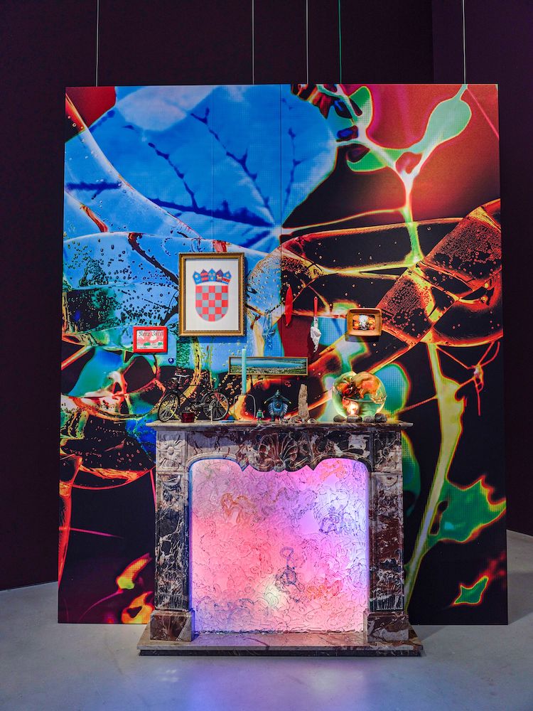 A marble mantelpiece is adorned with decorative items, in front of an abstract blue, green, and red backdrop.