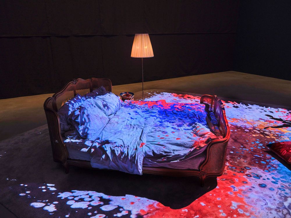 A bed and a lamp, bathed in the blue and red lights projected over them.