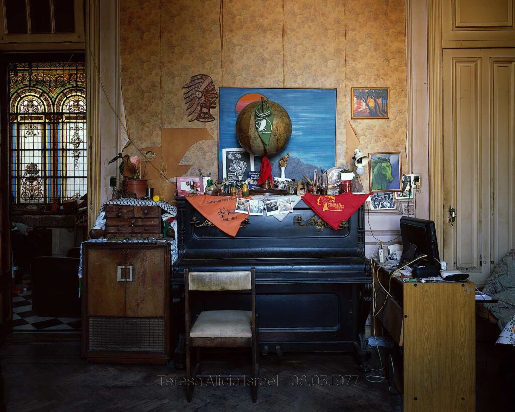 Colour photograph of a room featuring a piano, a globe, several trinkets along with a desk, the image says ‘Teresa Alicia Israel 08.03.1977’