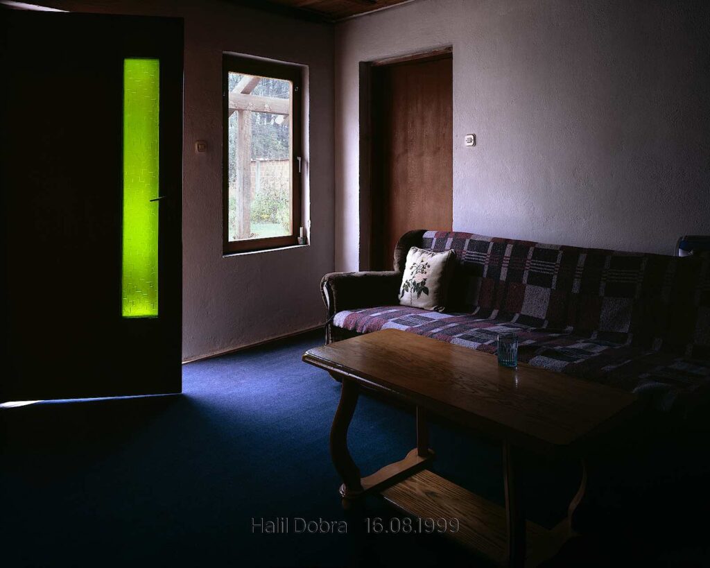 Colour photograph of a darkened room with a sofa, table and a door with green glass, the image states ‘Halil Dobra 16.08.1999’