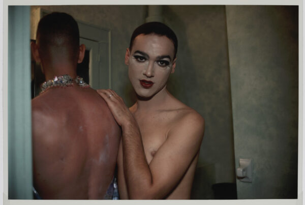 Portrait photograph of two men in a bathroom. One person looks towards the camera, wearing dramatic eye make up and dark red lipstick. Another man, wearing a dramatic necklace, is turned away from the camera. They are both shirtless.