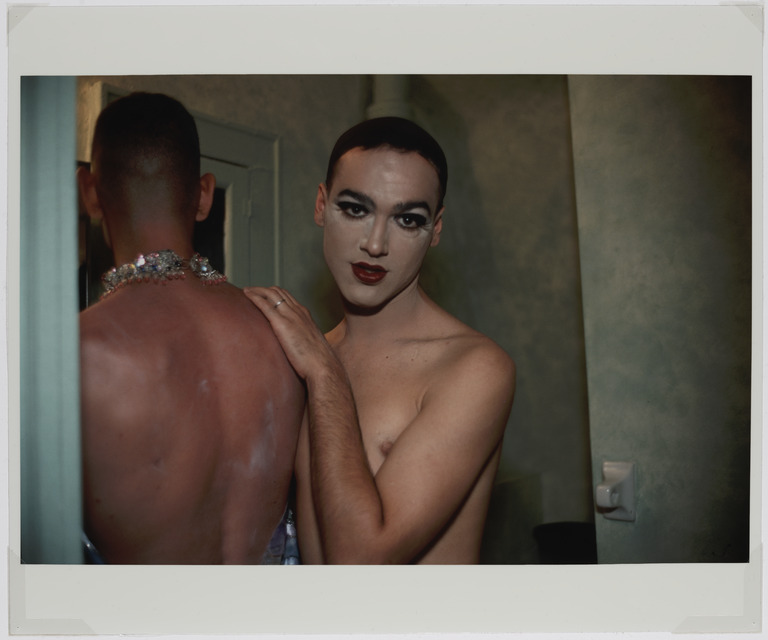 Portrait photograph of two men in a bathroom. One person looks towards the camera, wearing dramatic eye make up and dark red lipstick. Another man, wearing a dramatic necklace, is turned away from the camera. They are both shirtless.