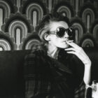 Woman smoking cigarette and drinking beer against backdrop of 1970s wallpaper