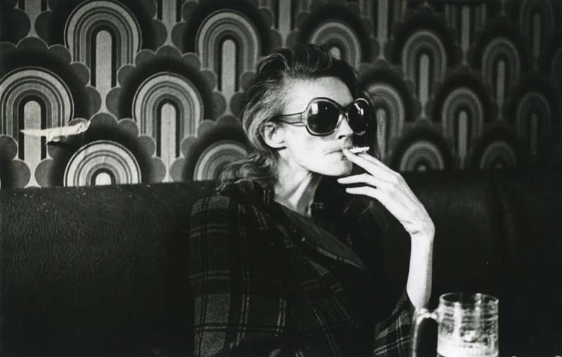 Woman smoking cigarette and drinking beer against backdrop of 1970s wallpaper