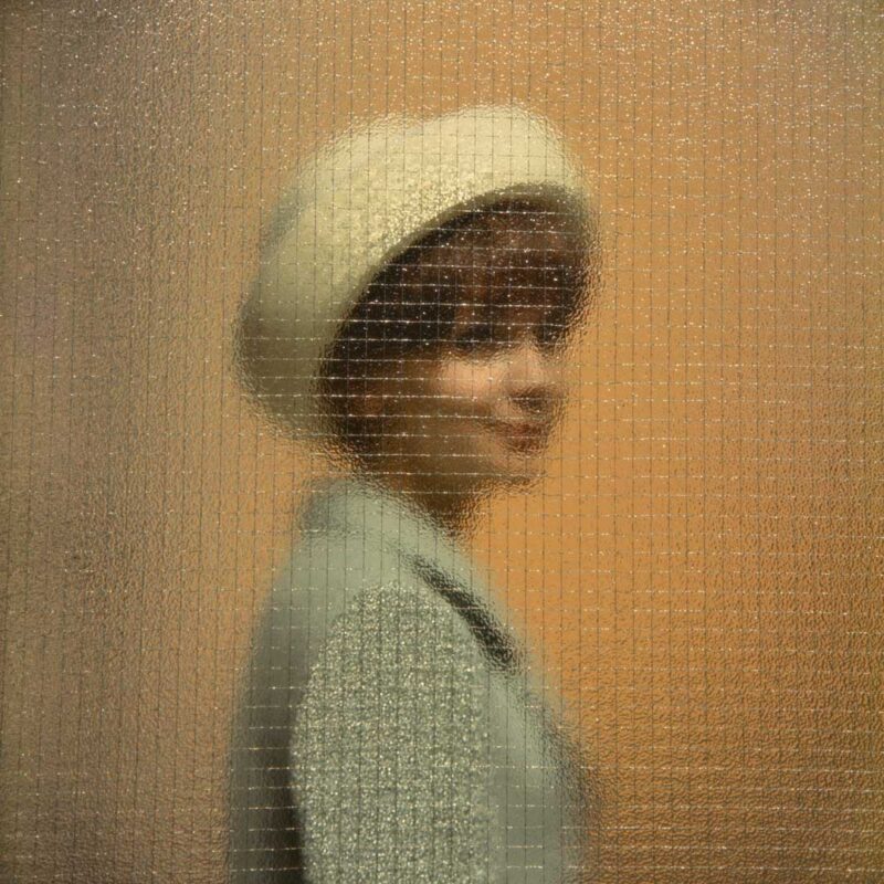 Woman in a green coat and a white hat behind a glass window