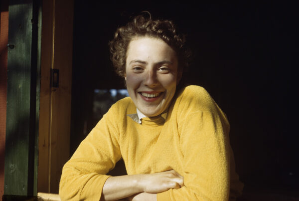 Woman with brown curly hair, smiling wearing a yellow jumper.